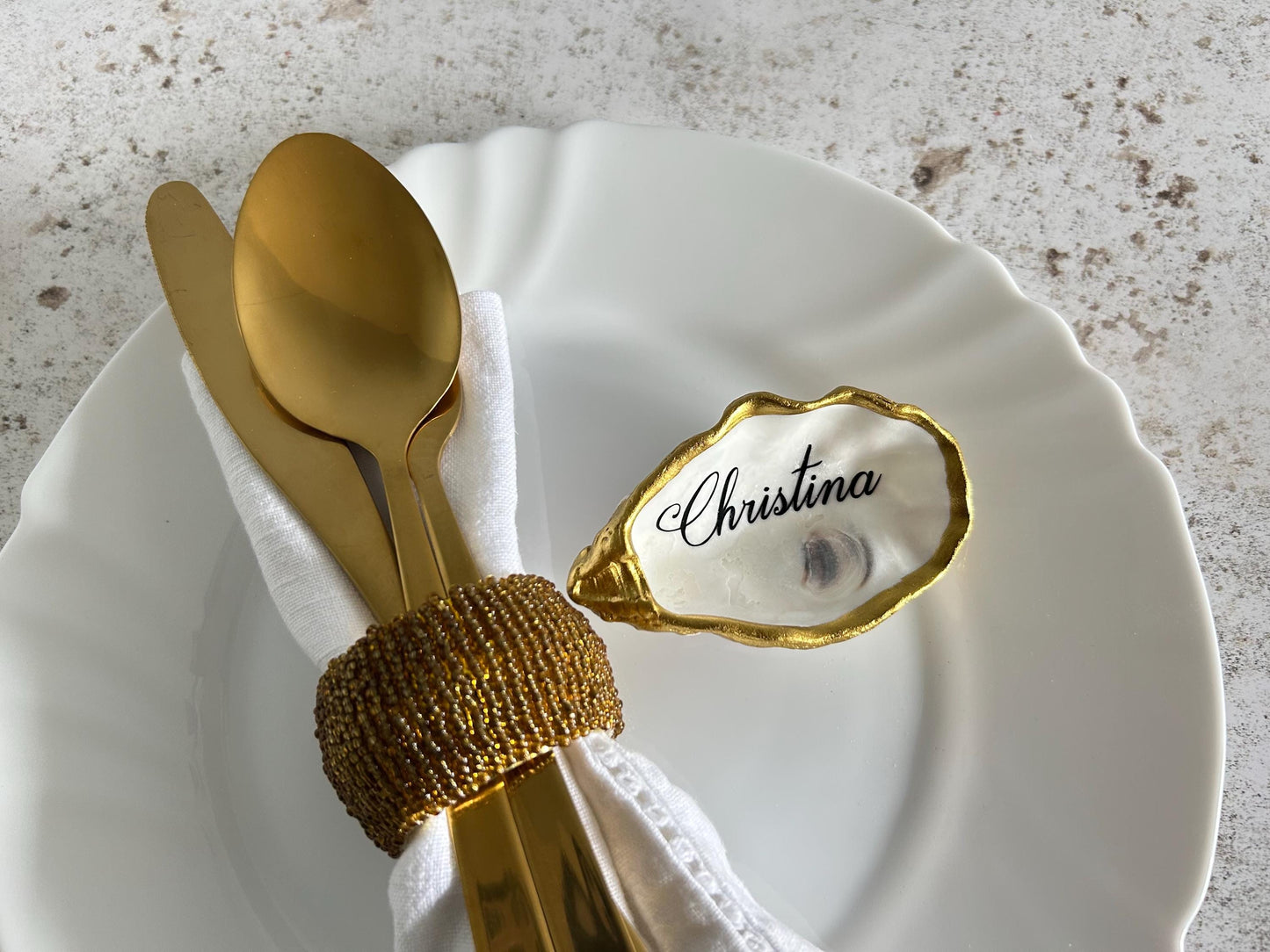 A gold oyster shell place card name with the name Christina written in black cursive. The place card holder is on a white plate with a gold napkin ring and gold silverware.