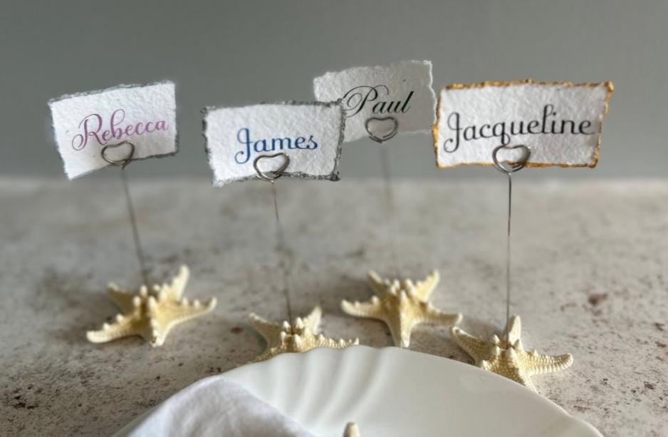 Place cards are available, they are hand torn seeded place cards with names written in calligraphy. The cards are held by silver wire place card holders with heart shaped tops adjoined on top of starfish