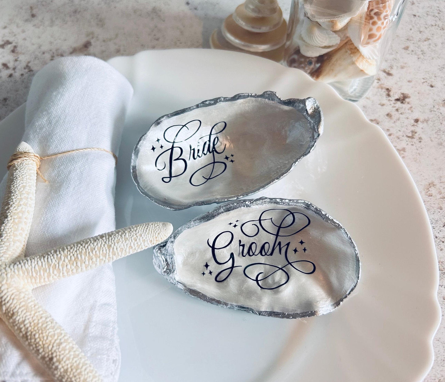 These silver oyster shells have black calligraphy. The shells say  'Bride' and  'Groom'.