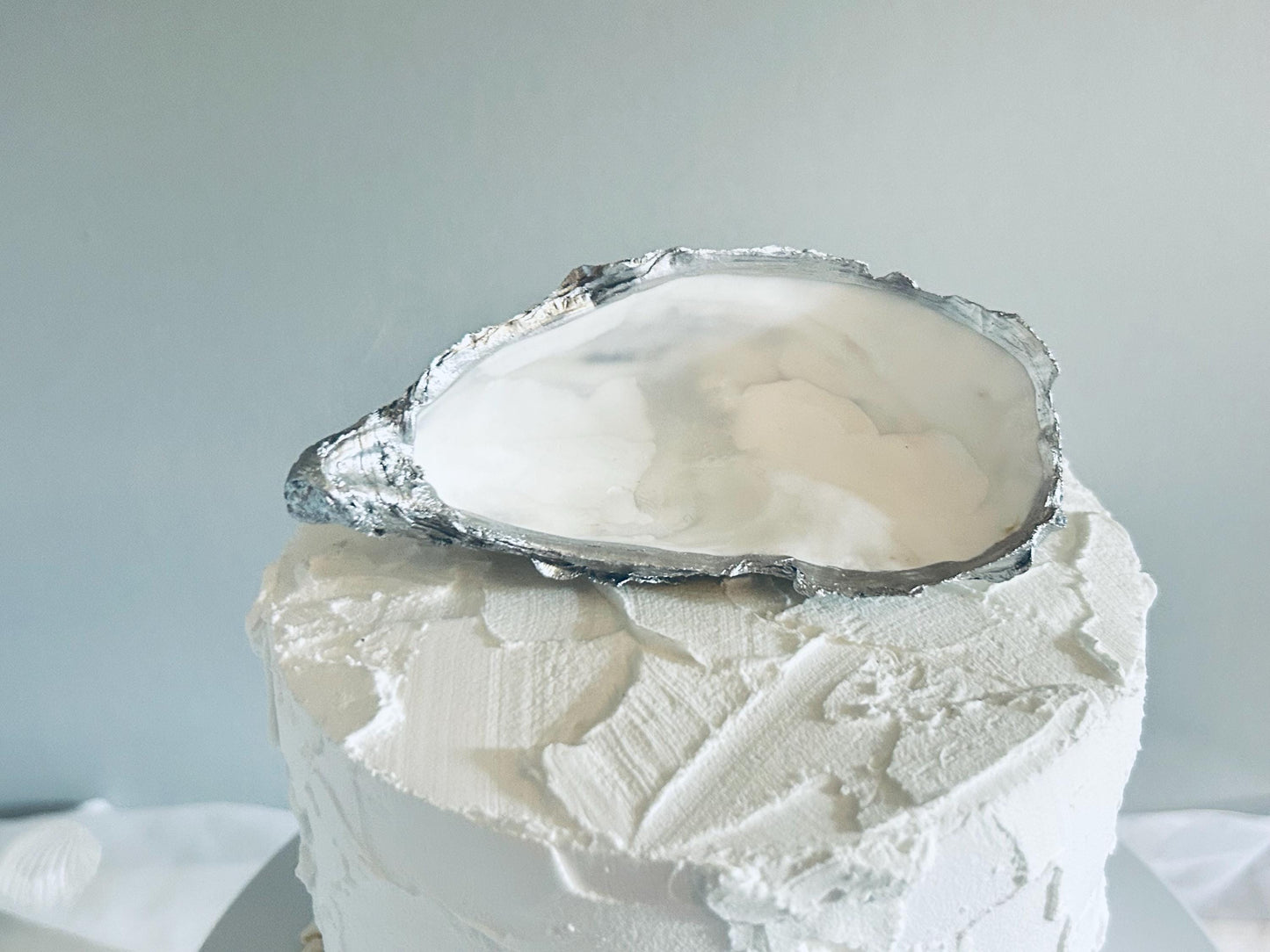 A white cake with textured buttercream frosting, topped with a large oyster shell with a silver rim. The shell is open, revealing a pearly interior which can be personalised . The cake sits on a silver cake stand.