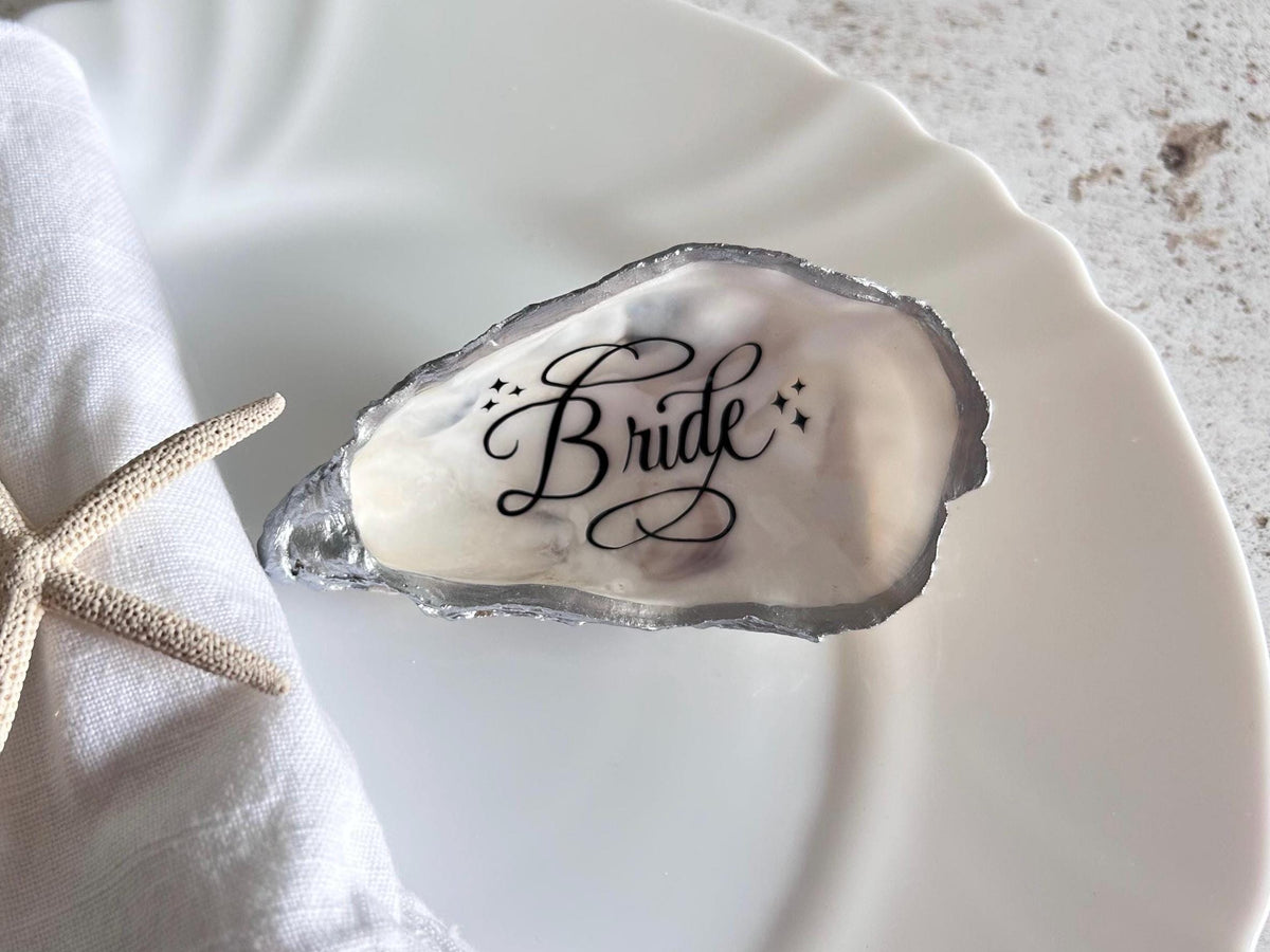 A white oyster shell with personalisation written in black calligraphy. The shell is natural on the inside and with bright silver metallic silver edges.