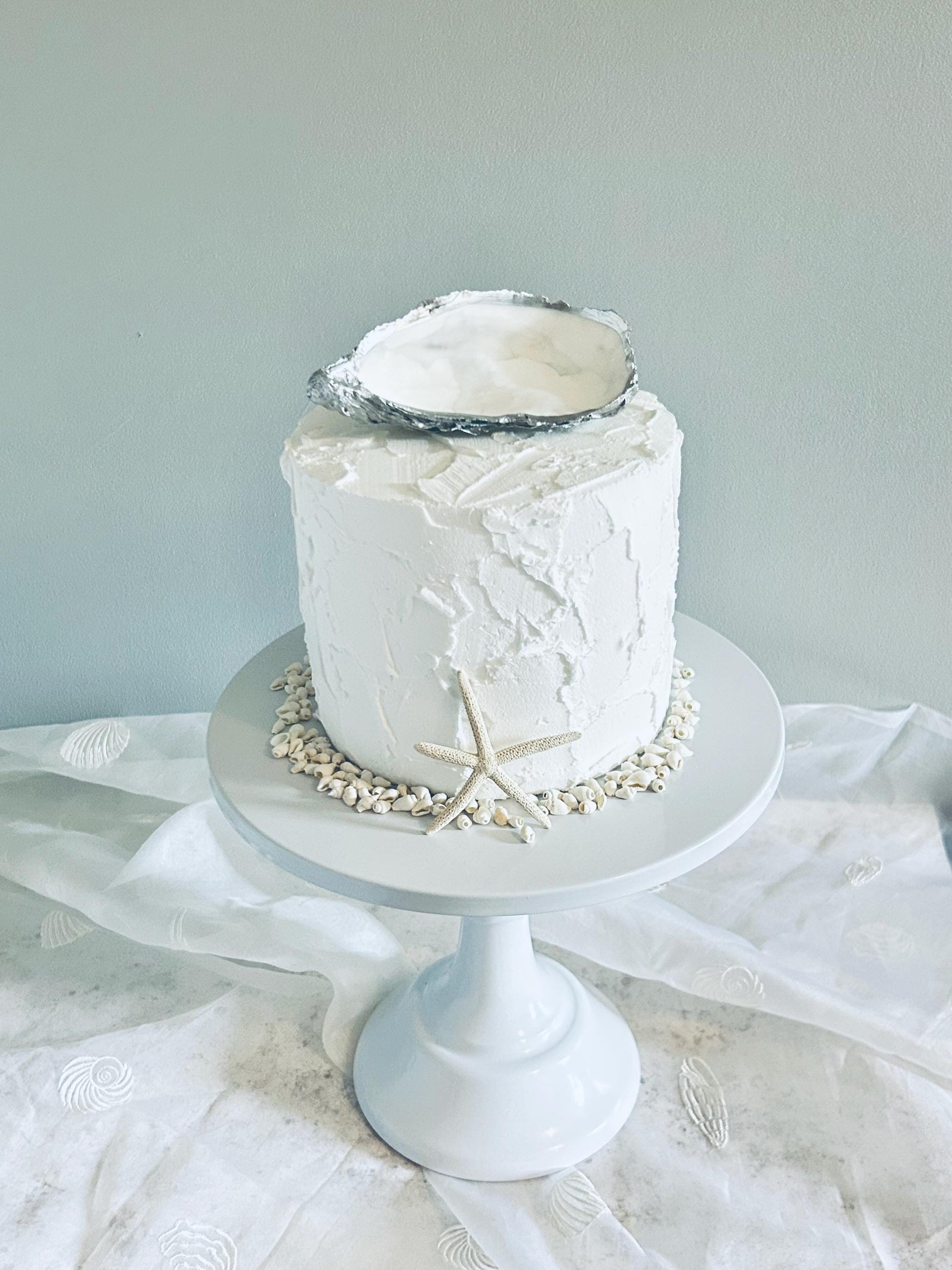 A oyster shell cake topper. The cake sits on a white pedestal cake stand, surrounded by small seashells and a starfish.