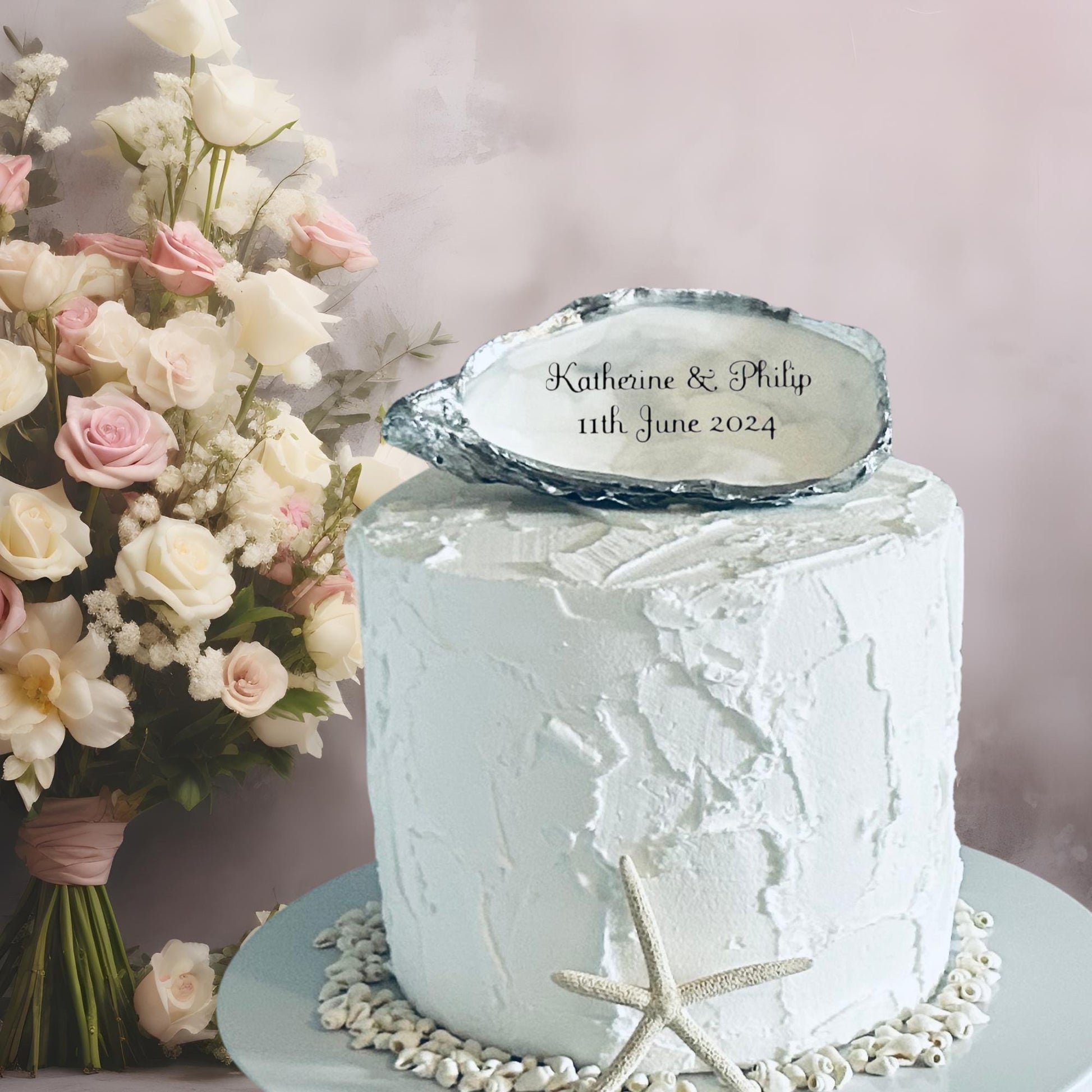 A white wedding cake with textured frosting, adorned with an oyster shell inscribed with a personalisation of your choice. A bouquet of pink and white roses and a starfish decorate the cake, suitable for a wedding.