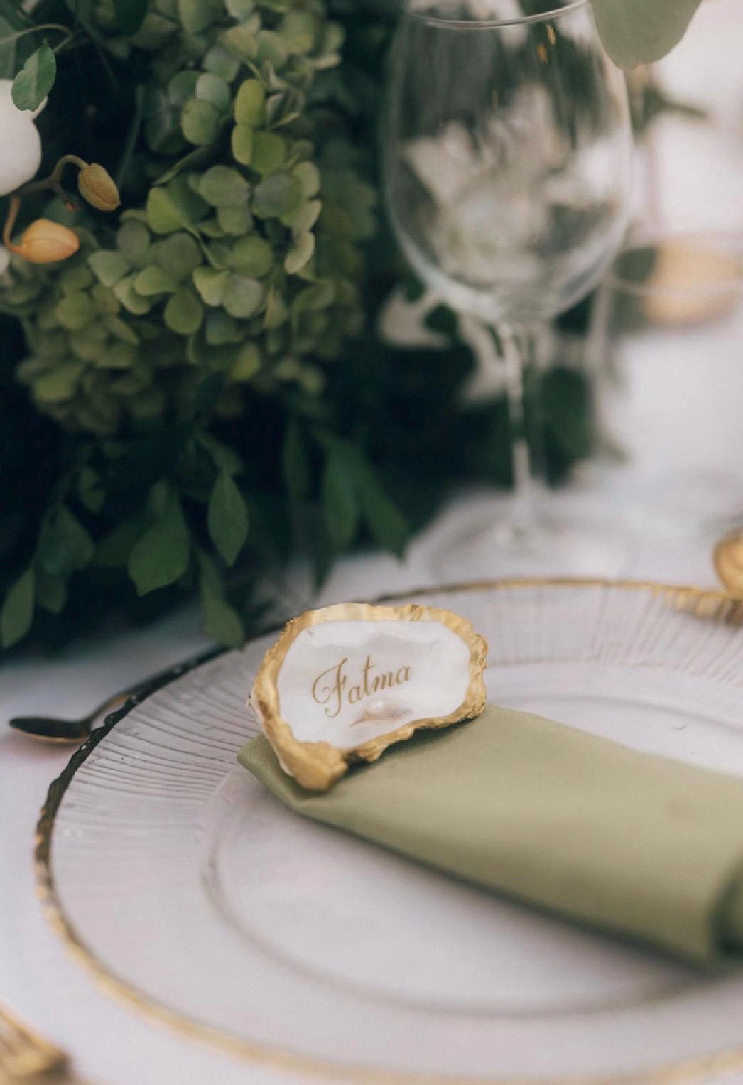 A table setting with a white plate with a gold rim, a folded green napkin, and an oyster shell name card. The name is written in gold script on the shell. Green foliage and a wine glass are in the background.