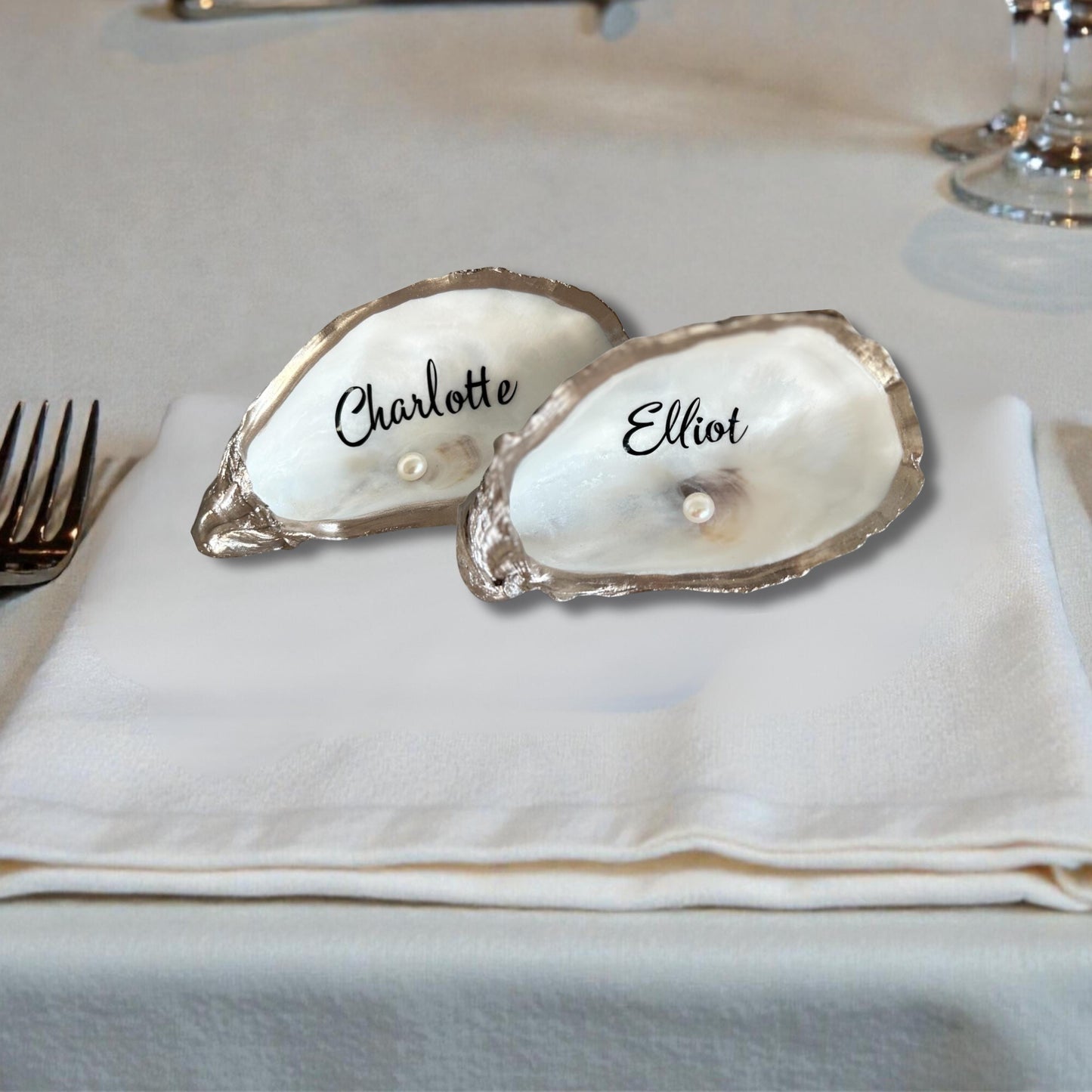 Two oyster shell place cards with champagne gold edging, each inscribed with a name in black script. Each features a small pearl. The shells are placed on a white napkin.