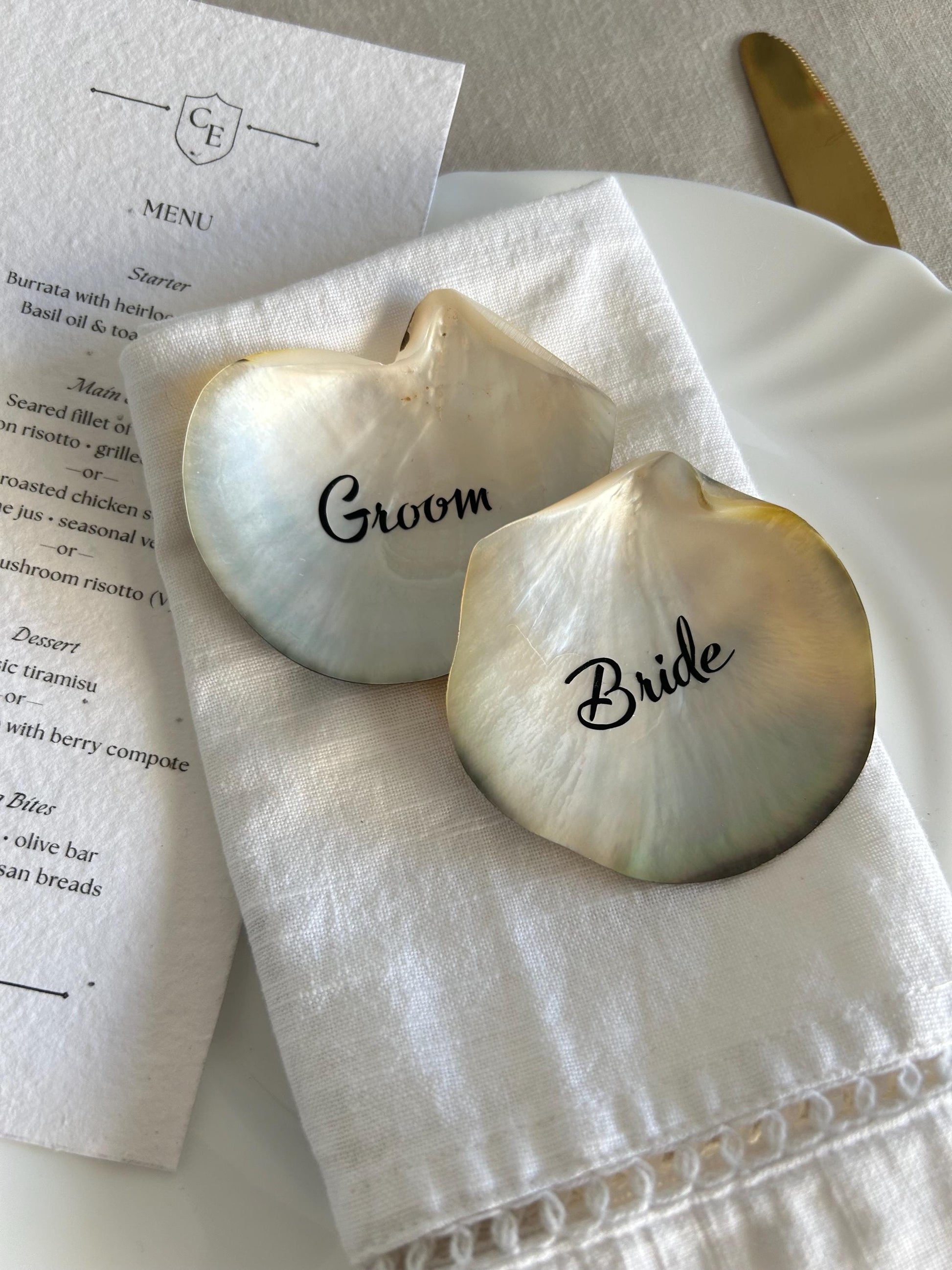 Two iridescent seashell place cards, labelled "Groom" and "Bride", sit on a white linen napkin.