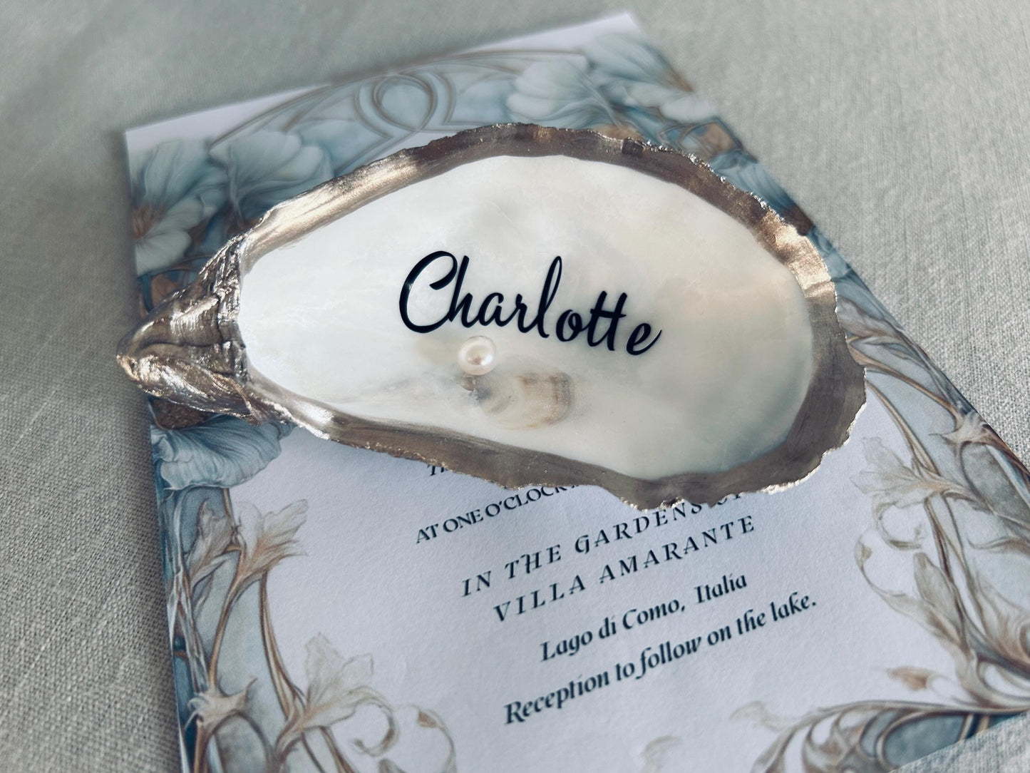 An oyster shell place card holder with the name "Charlotte" in black script, a small pearl, and a wedding invitation with floral details. The invitation is for a wedding in Italy.