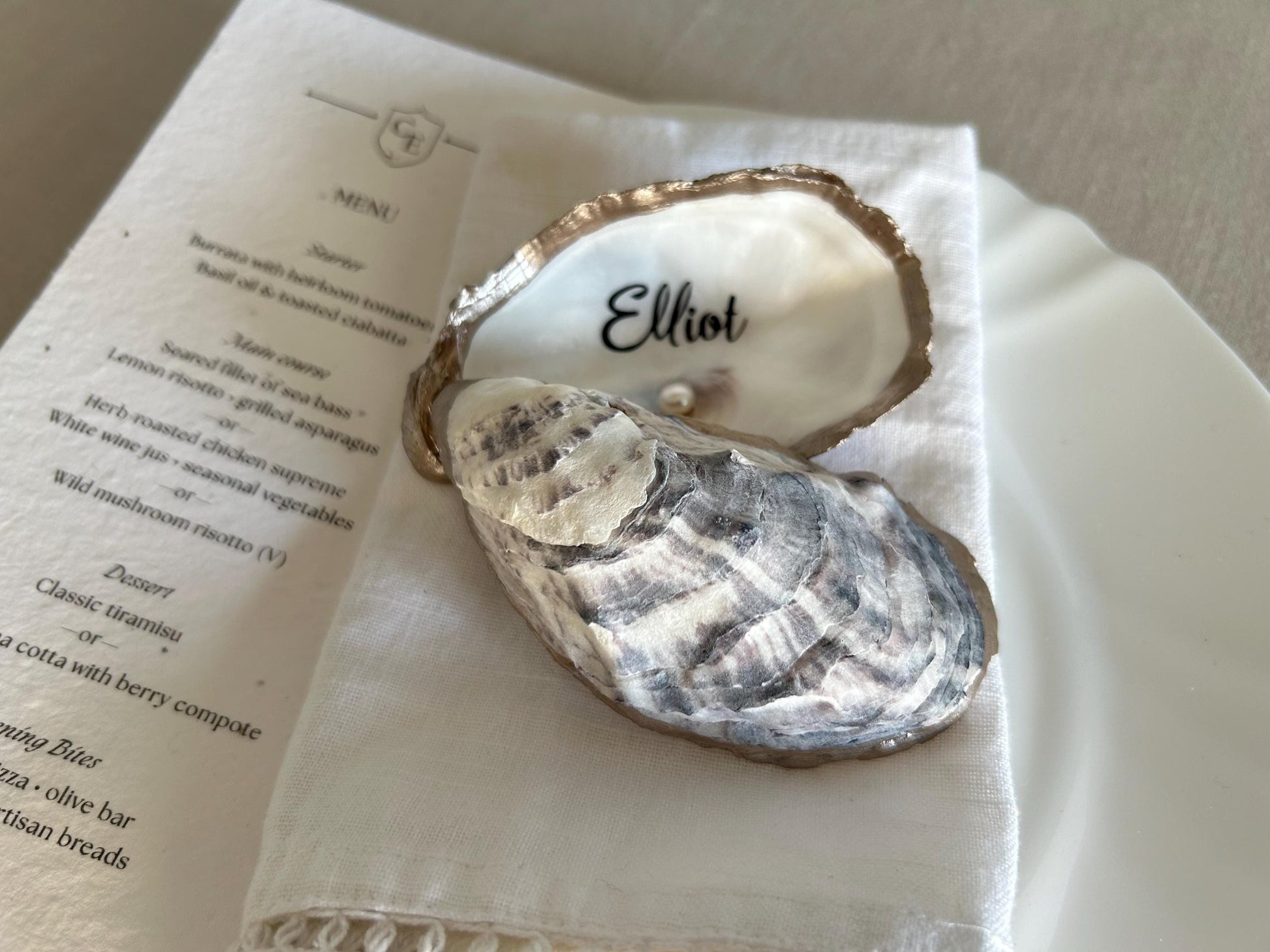 An open oyster shell with the name written inside, resting on a white napkin. The shell is gray and white on the underside