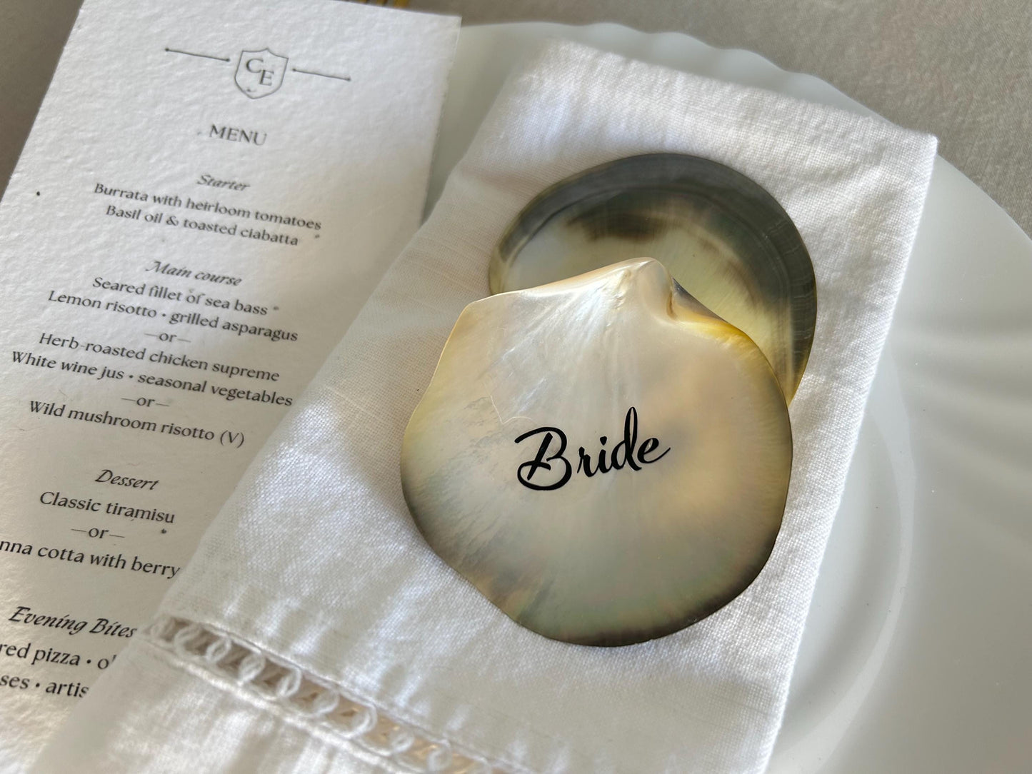 Two iridescent seashell place cards, one with the word "Bride", rest on a white linen napkin.