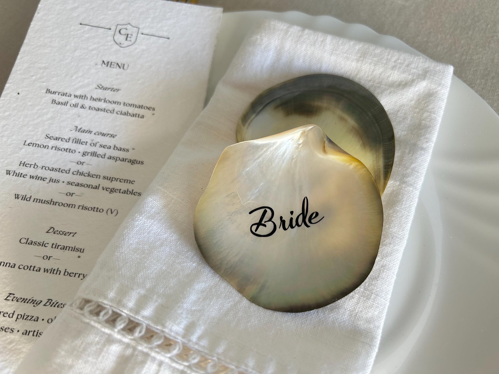 Two iridescent seashell place cards, one with the word "Bride", rest on a white linen napkin.