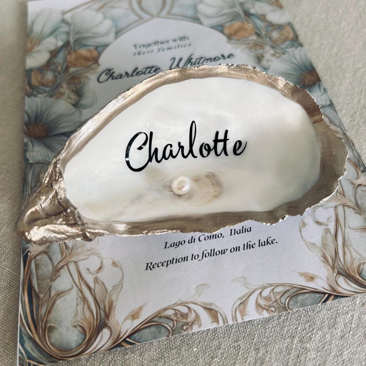 A white oyster shell and the name in black script. A small pearl is inside the shell which is optional
