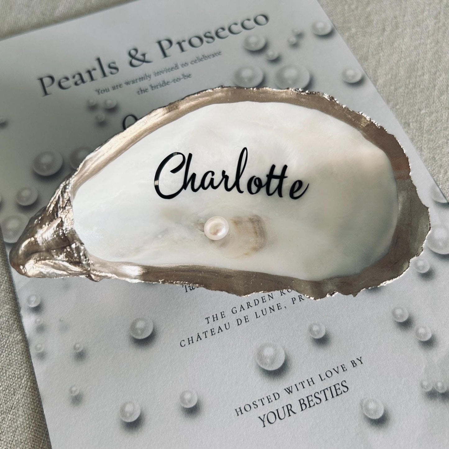 a decorative oyster shell with a pearl and the name  written on it. The shell sits on a card with the text "Pearls & Prosecco" and pearl illustrations, suitable for a wedding or event.